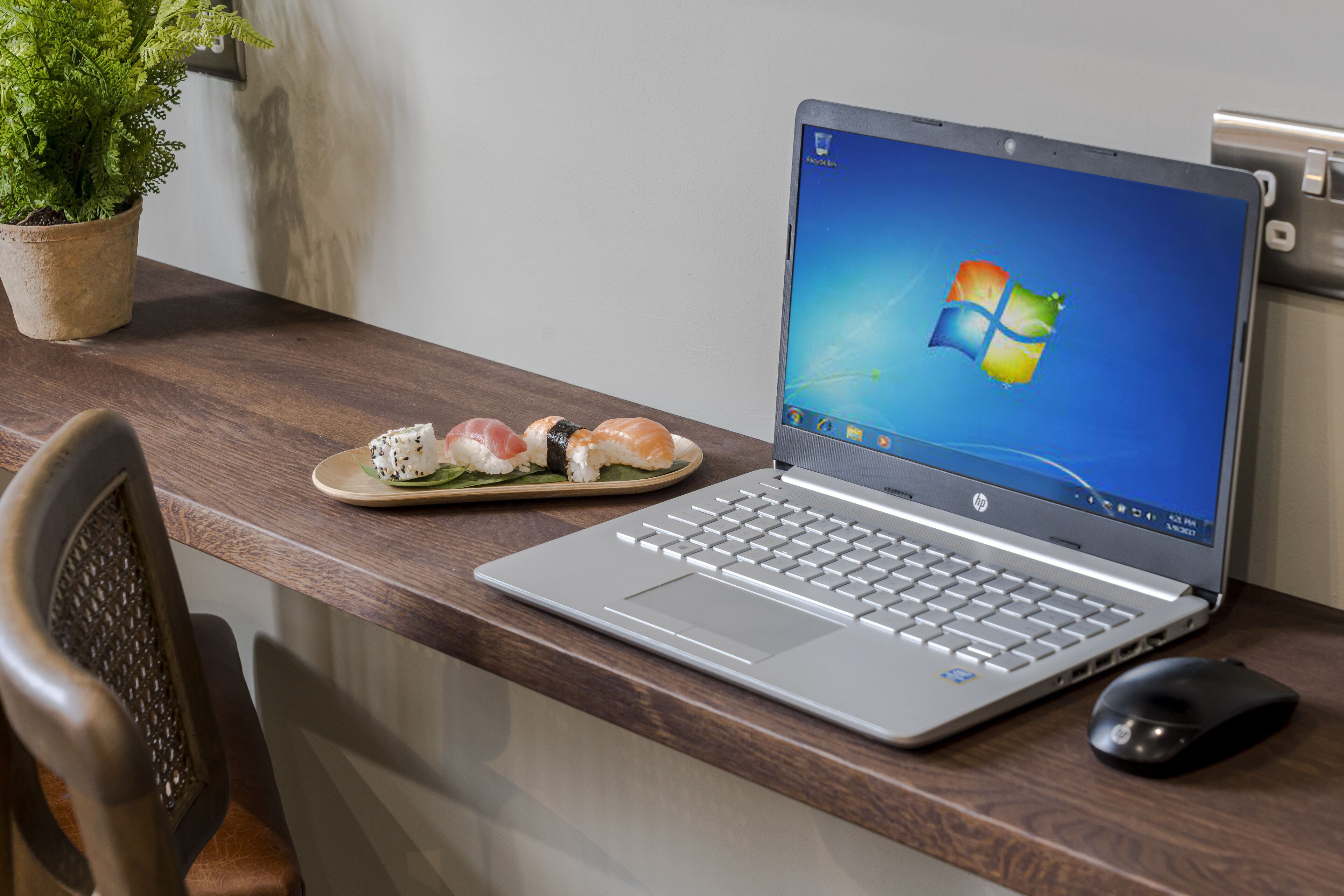 A laptop on a wooden desk with a plate of sushi and a potted plant.