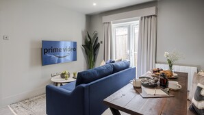 A modern living room with a blue sofa, a dining table with breakfast items, a coffee maker, and a TV displaying 'prime video'. - Your Apartment Tooting (London)