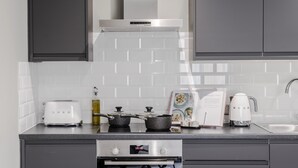 A modern kitchen with grey cabinets, white tiled backsplash, a stainless steel range hood, a toaster, a kettle, a frying pan, and a cookbook. - Your Apartment Tooting (London)
