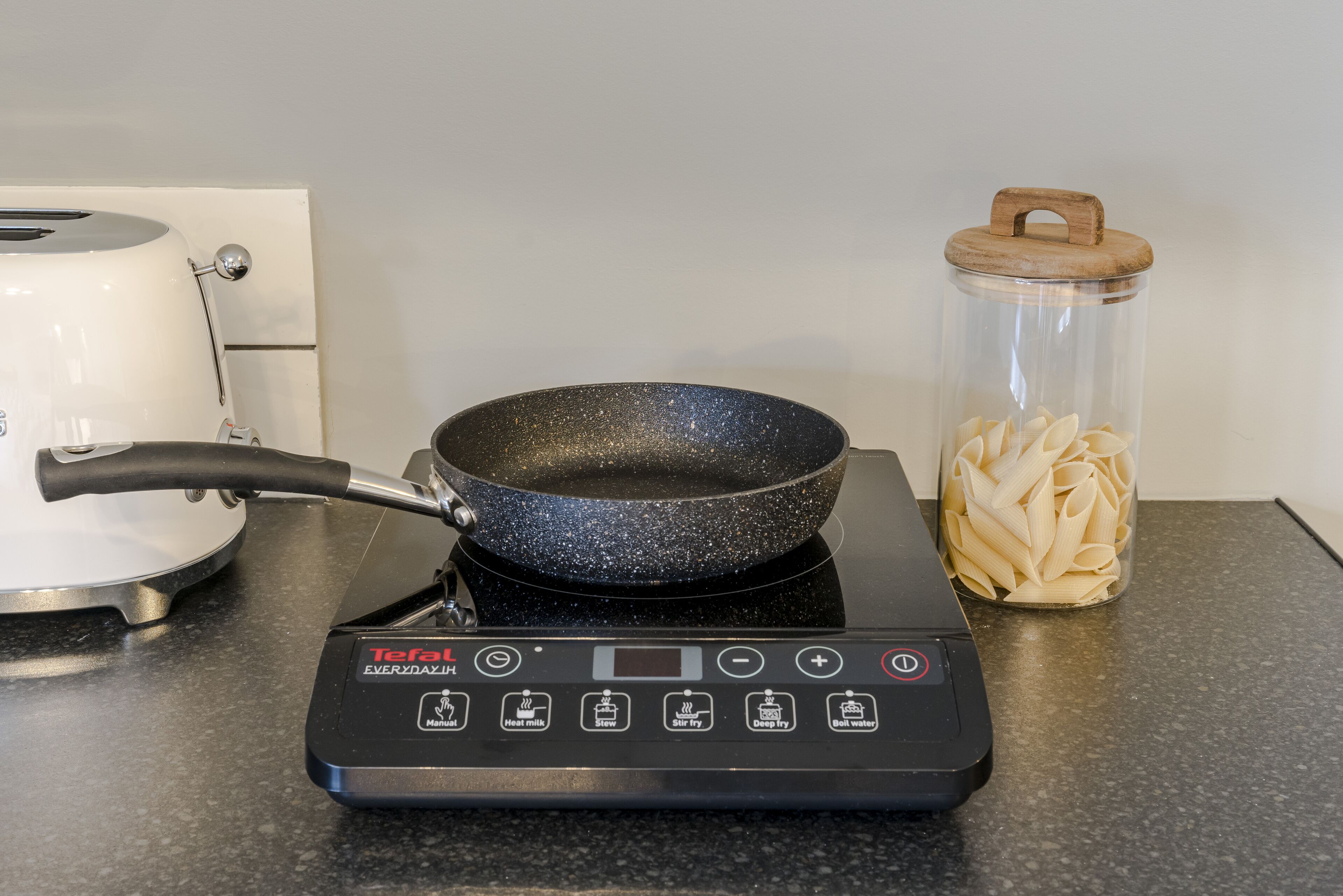 A kitchen with a black electric stove, a frying pan, a white kettle, and a glass jar with pasta.