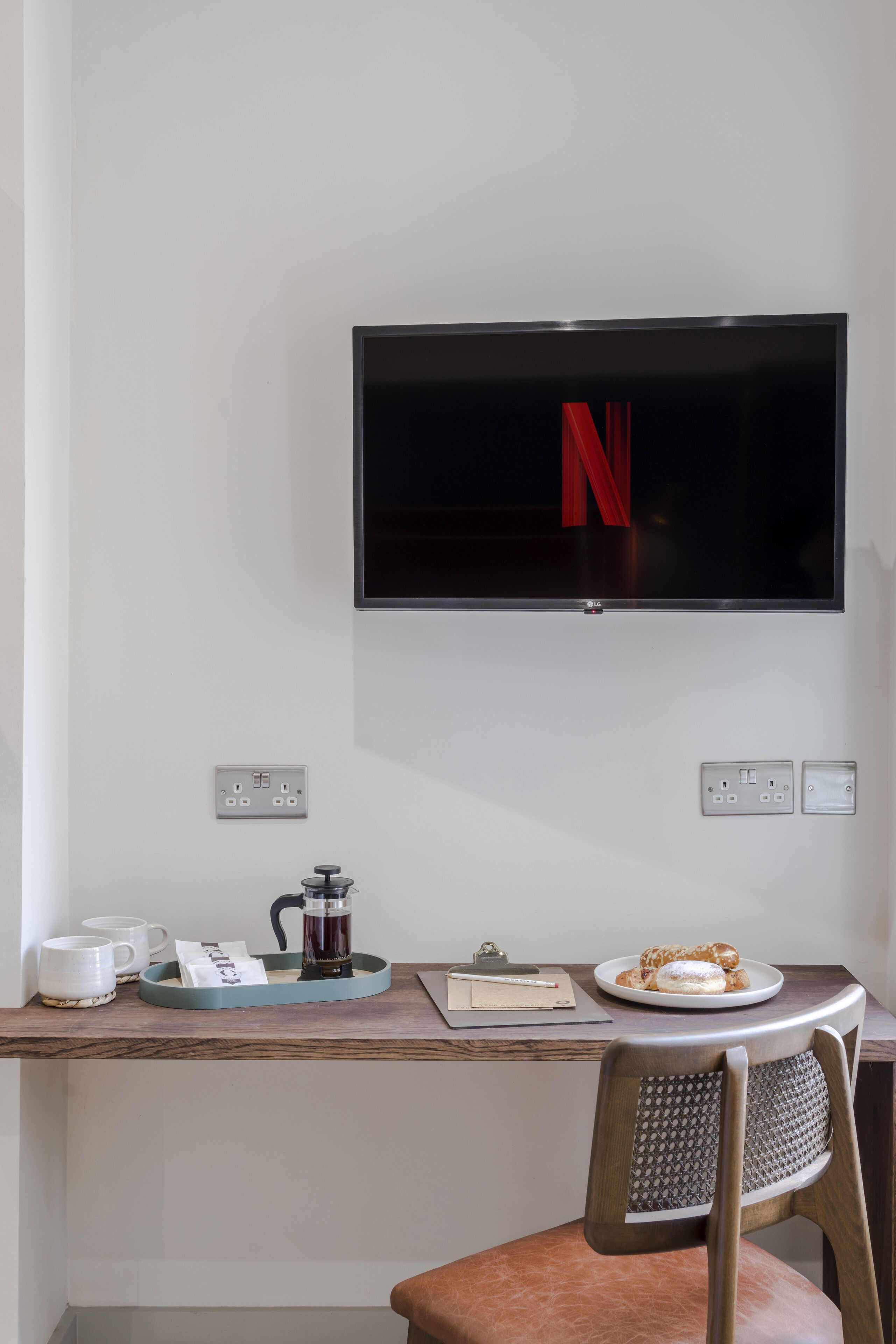 A modern room with a flat-screen TV mounted on the wall, a wooden desk with a coffee maker, cups, and a plate of pastries.