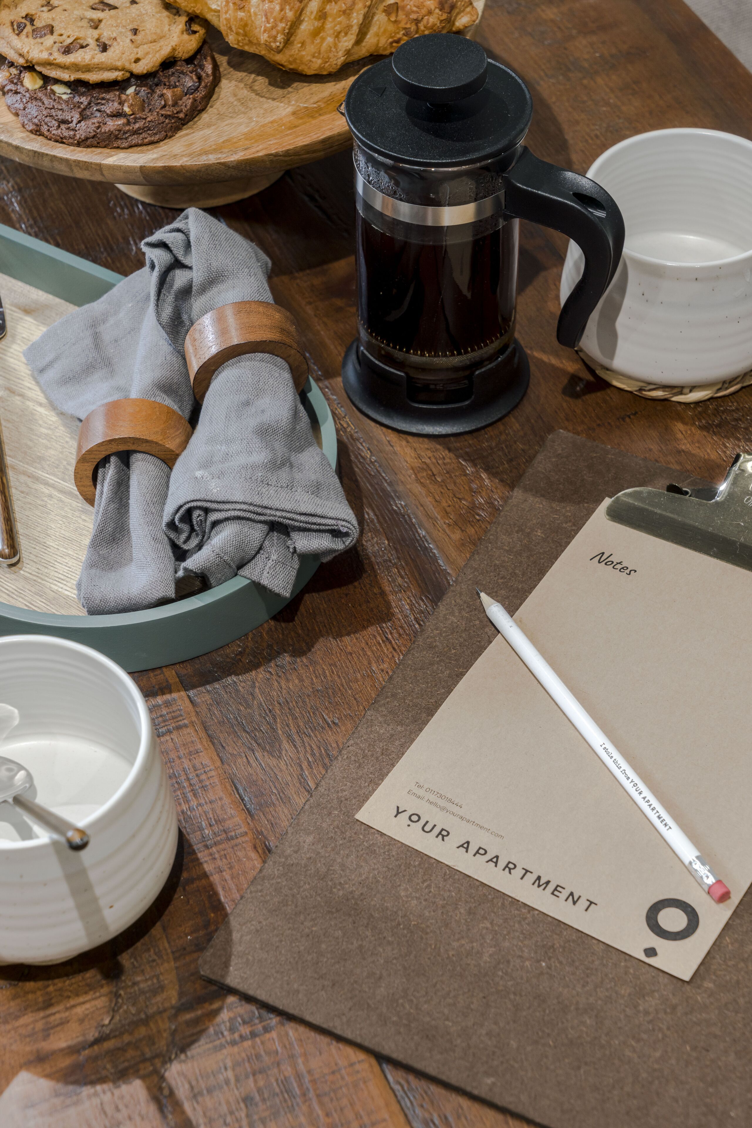 A wooden table with a coffee maker, a cup, a plate with cookies, a notebook, and a pen.