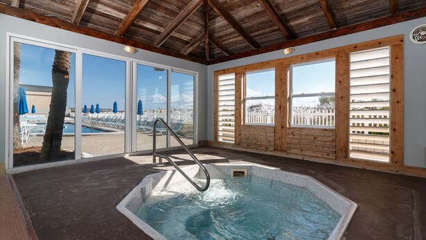 A hot tub with a wooden deck and a view of a beach through large windows.