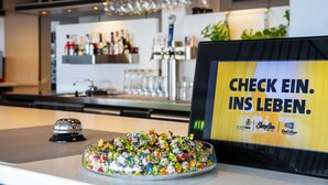 A bar area with a variety of bottles and glasses, a bowl of colorful candies, and a sign with text. - [BEE]VILLAGE MESSEHOTEL HANNOVER-LAATZEN. (Laatzen)