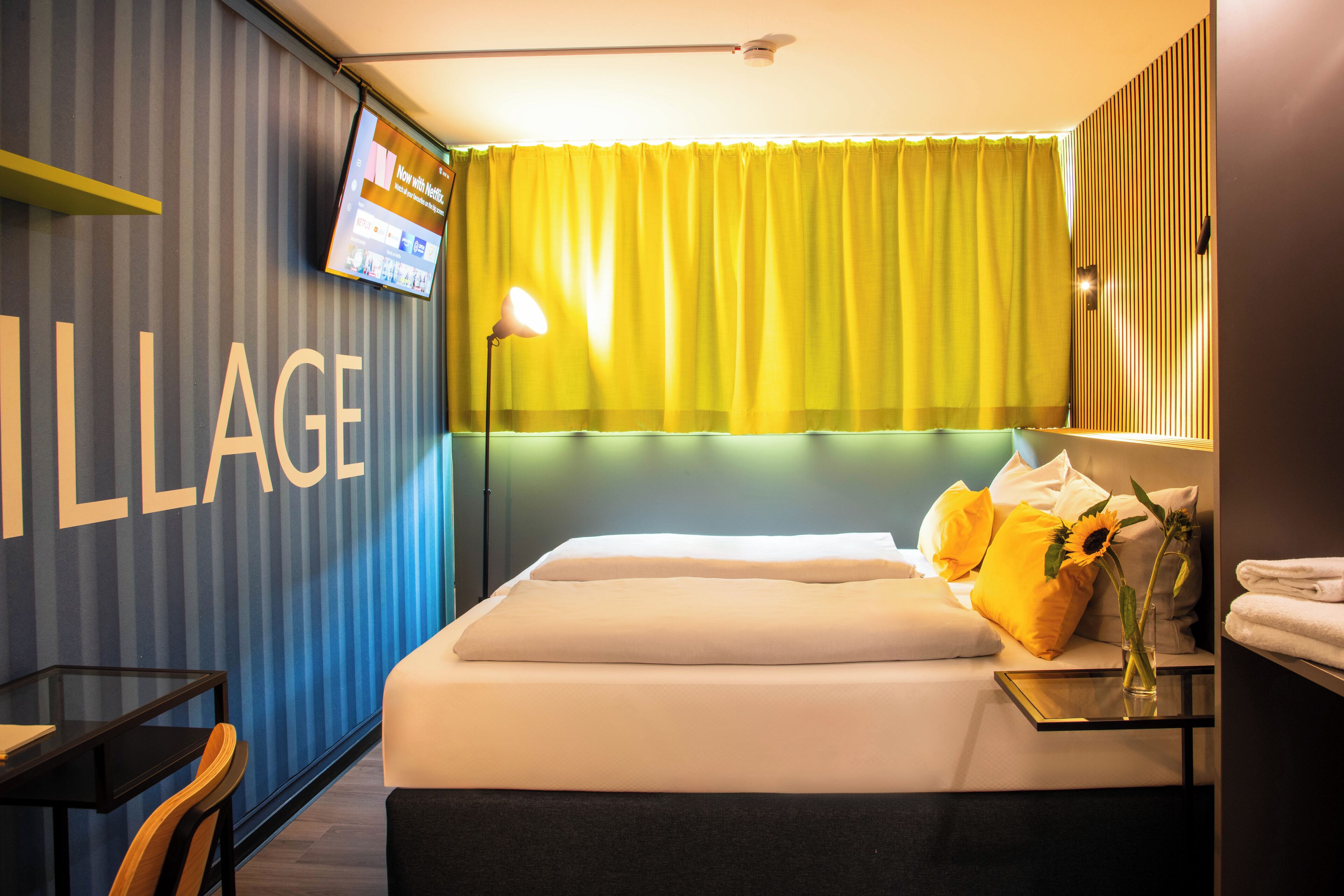Une chambre d’hôtel avec un lit, un téléviseur fixé au mur, une table de chevet avec une lampe et une fenêtre avec des rideaux jaunes.