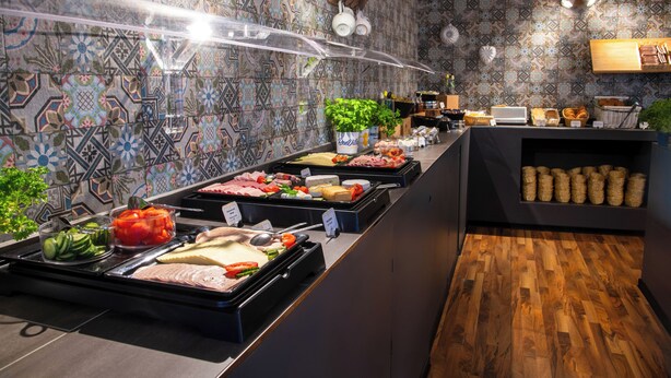 A buffet with various food items, a patterned wall, and a wooden floor. - [BEE]VILLAGE MESSEHOTEL HANNOVER-LAATZEN. (Laatzen)