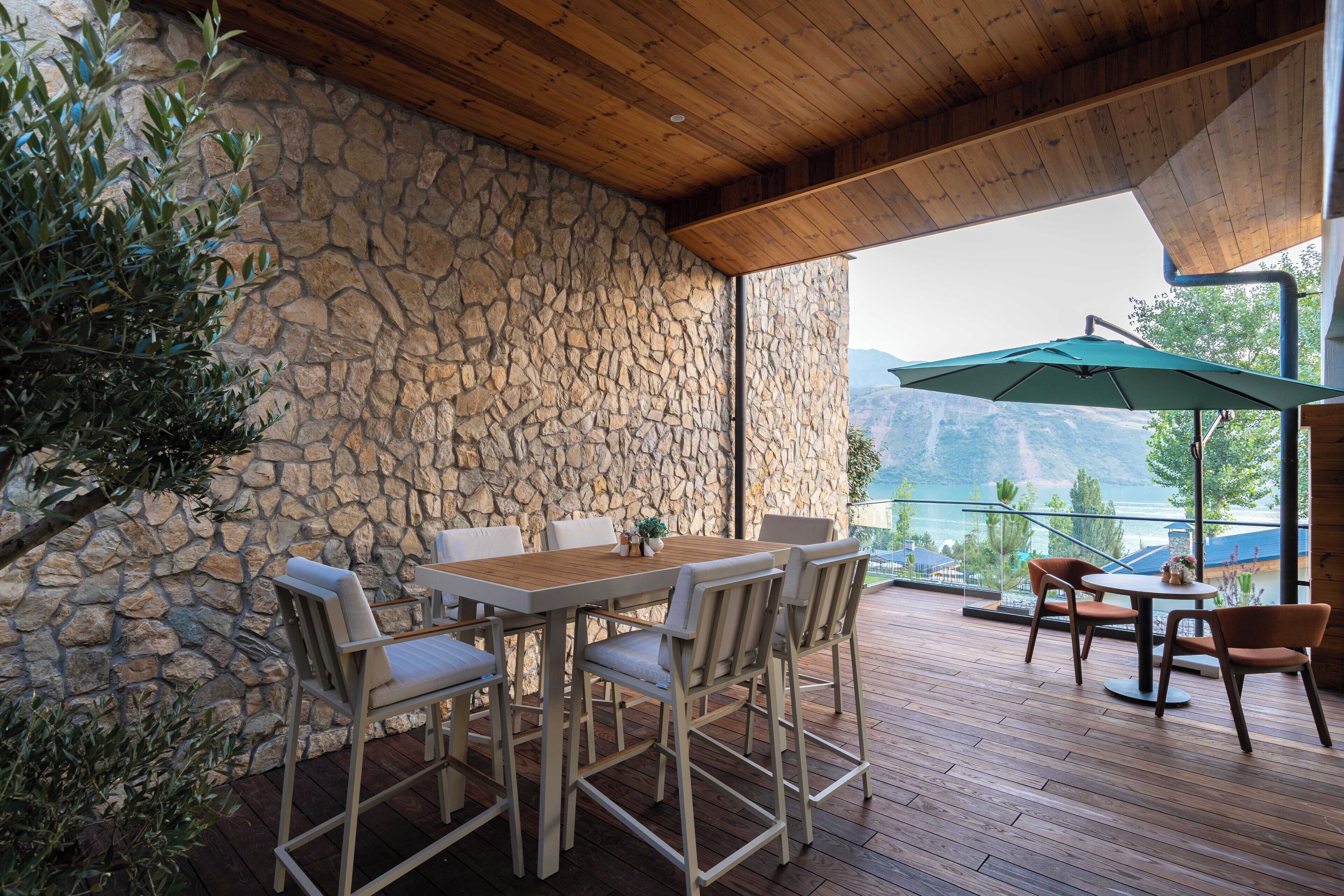 Zona pranzo all'aperto con parete in pietra, terrazza in legno e vista sulle montagne.