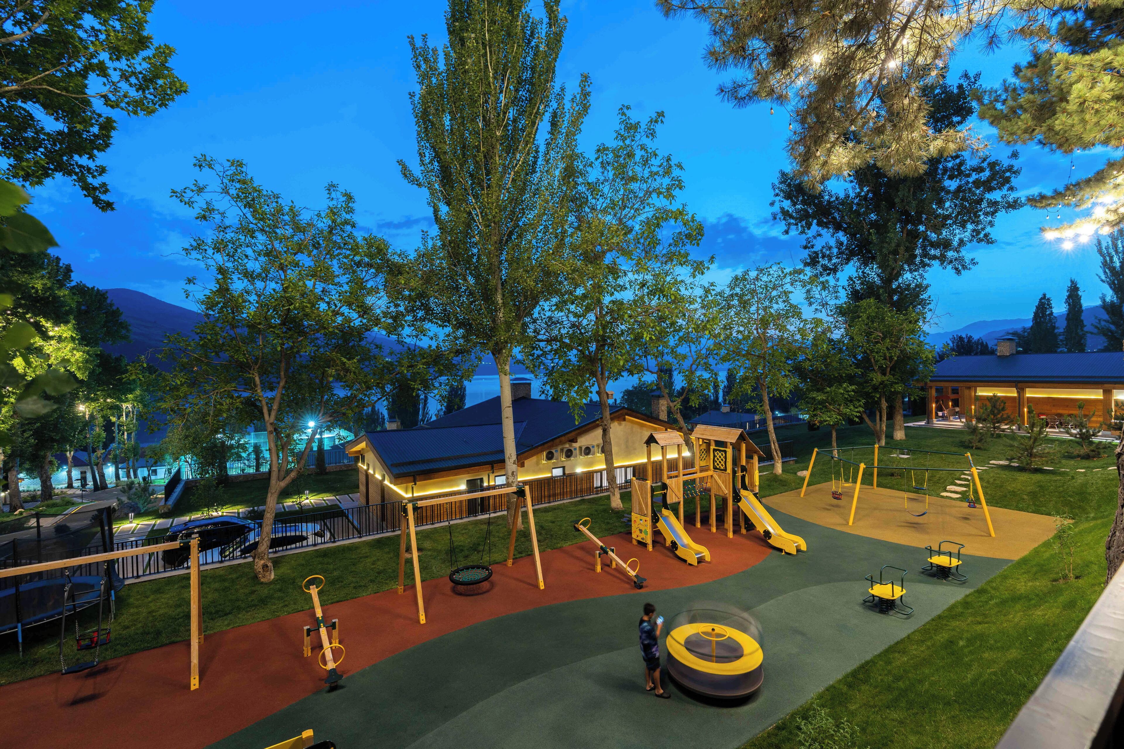 A playground with swings, slides, and climbing structures, surrounded by trees and a building in the background.