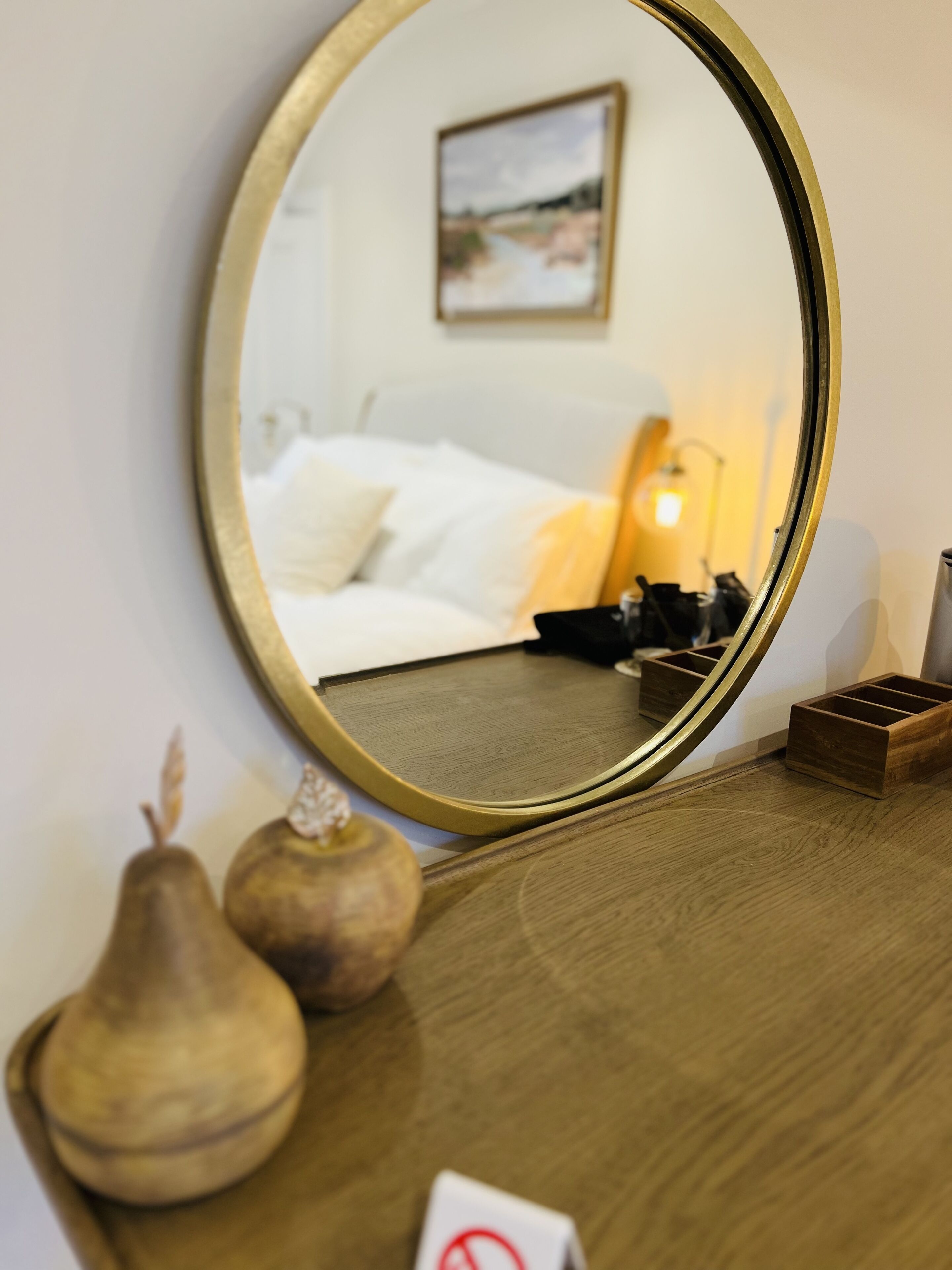 A round mirror on a wooden dresser reflecting a bed, pillows, a lamp, and a small wooden box.
