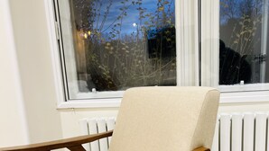 A modern armchair with wooden armrests and a beige fabric seat, positioned in front of a window with a view of trees and a clear sky. - St Duthus Villa Guesthouse (Tain)