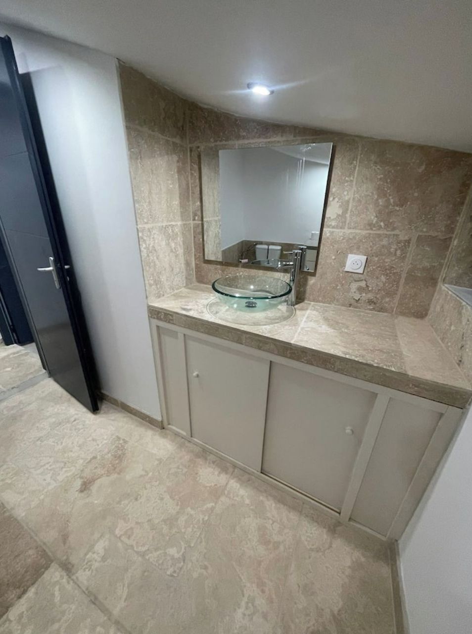 Une salle de bain avec un lavabo en verre, un miroir au-dessus, et des murs recouverts de carreaux beiges.