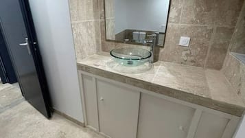Une salle de bain avec un lavabo en verre, un miroir au-dessus, et des murs recouverts de carreaux beiges.