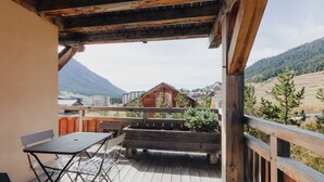 Une terrasse en bois avec une table et des chaises, offrant une vue sur un paysage montagneux avec des bâtiments visibles au loin.