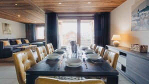Un coin repas avec une table en bois dressée pour six personnes, entourée de fauteuils en cuir, et offrant une vue sur une maison à travers une grande fenêtre.