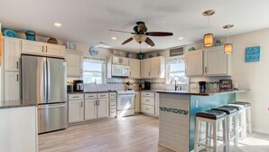 A modern kitchen with a stainless steel refrigerator, white cabinets, a central island with bar stools, and a ceiling fan. - ☀️Atlantic Views- Beautiful 8BR Home with Private Pool (Kure Beach)
