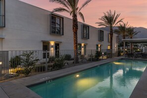 A swimming pool with a modern building in the background, a fenced area, and palm trees. - House of Light - Bright Modern 4BR Townhome (Palm Springs)