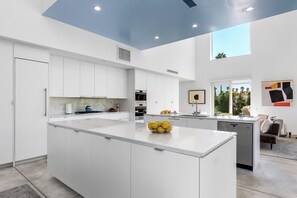 A modern kitchen with white cabinetry, a central island, and a large window providing a view of the outdoors. - House of Light - Bright Modern 4BR Townhome (Palm Springs)