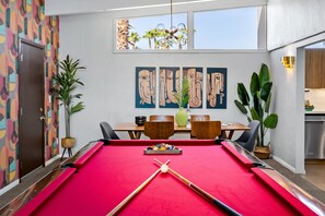Une table de billard recouverte d’un feutre rouge, un queue de billard et un jeu de billes. On y trouve également un coin repas avec des chaises et une table en bois, une plante et des œuvres d’art accrochées au mur.