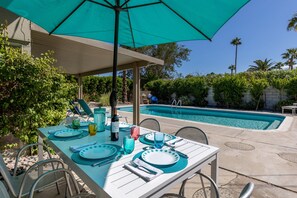 Un espacio exterior para comer con una mesa dispuesta para cuatro, una piscina al fondo y un porche cubierto.