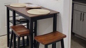 Zona pranzo con un tavolo in legno scuro e due sgabelli, un quadro con bottiglie di vino sulla parete, e una porta bianca.