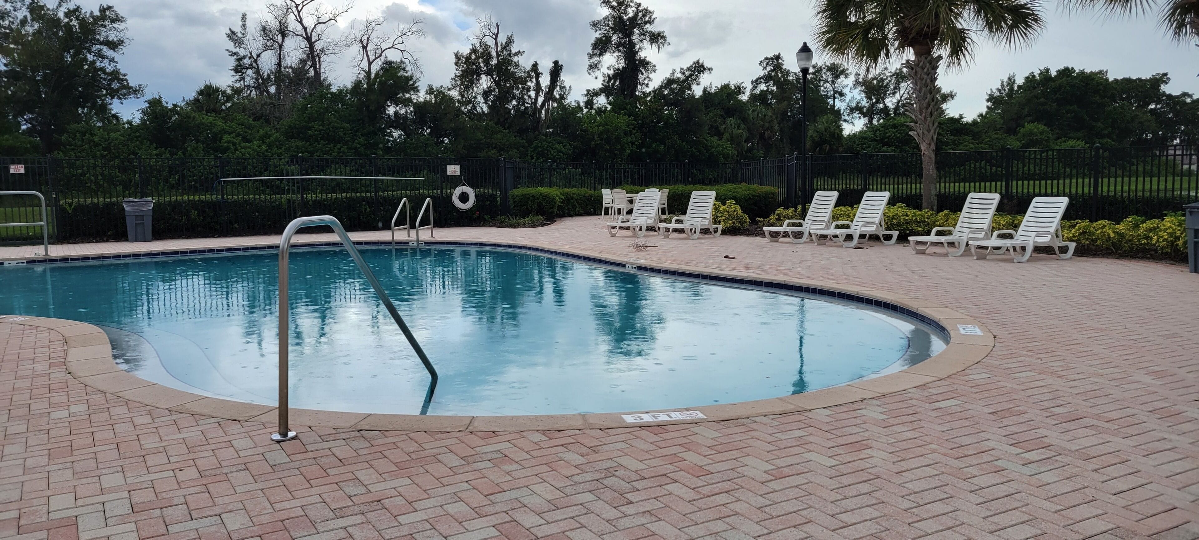 A swimming pool with a brick deck, a ladder, and white lounge chairs.