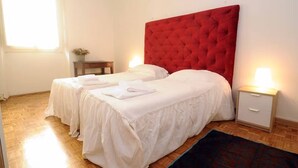 Hotellihuone, jossa on kaksi sänkyä, punainen pääty, valkoiset vuodevaatteet, pieni yöpöytä lampun kanssa ja ikkuna, jossa on verhot.