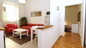 Ein gemütlicher Wohnraum mit einem roten Sofa, weißen Sesseln, einem kleinen Tisch und Holzboden.