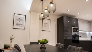 A modern dining area with a dark wooden table, grey upholstered chairs, and a bowl of fruit. - Exquisite 6 Bedroom Luxury Home (Manchester)
