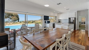 A modern kitchen with large windows, a dining area with a wooden table and white chairs, and a view of the ocean. - Encounter Blue Victor Harbor Retreat: Secluded Pool (Waitpinga)