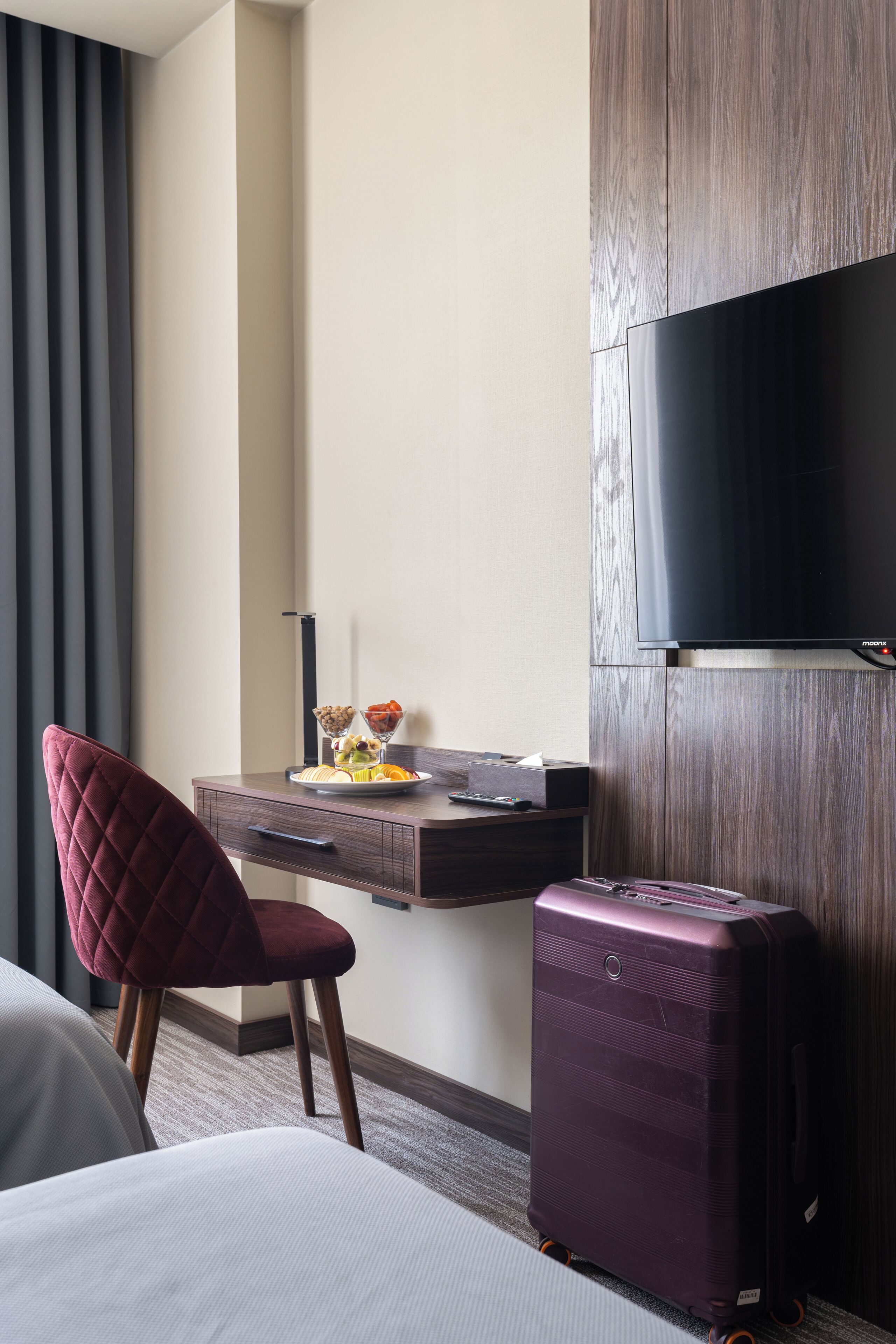 A hotel room with a bed, a desk with a flat-screen TV, a chair, a suitcase, and a window with blinds.