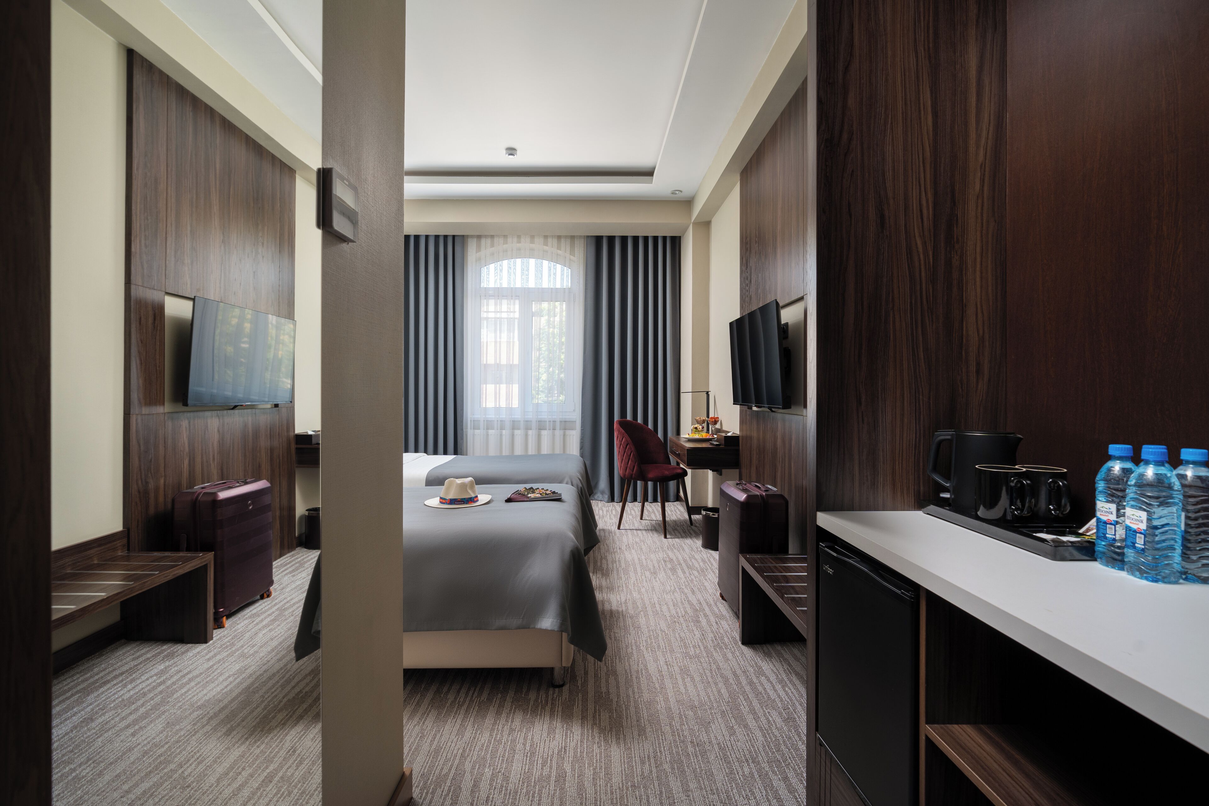 A hotel room with a bed, desk, chair, TV, and a minibar.
