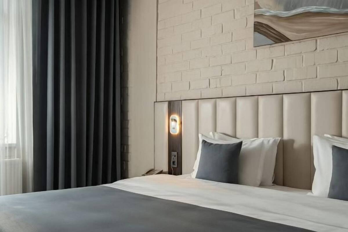 Ein Hotelzimmer mit einem großen Bett, einem Kopfteil mit einer Leuchte, einem Fenster mit Vorhängen und einer Ziegelwand.