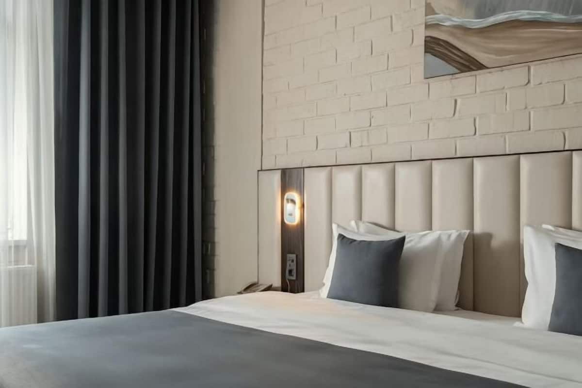 A hotel room with a large bed, a headboard with a light fixture, a window with curtains, and a brick wall.