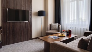 A modern hotel room with a flat-screen TV mounted on a wooden panel wall, a sofa, a coffee table with a magazine and snacks, and a floor lamp. - StayInn City Center (Tashkent)
