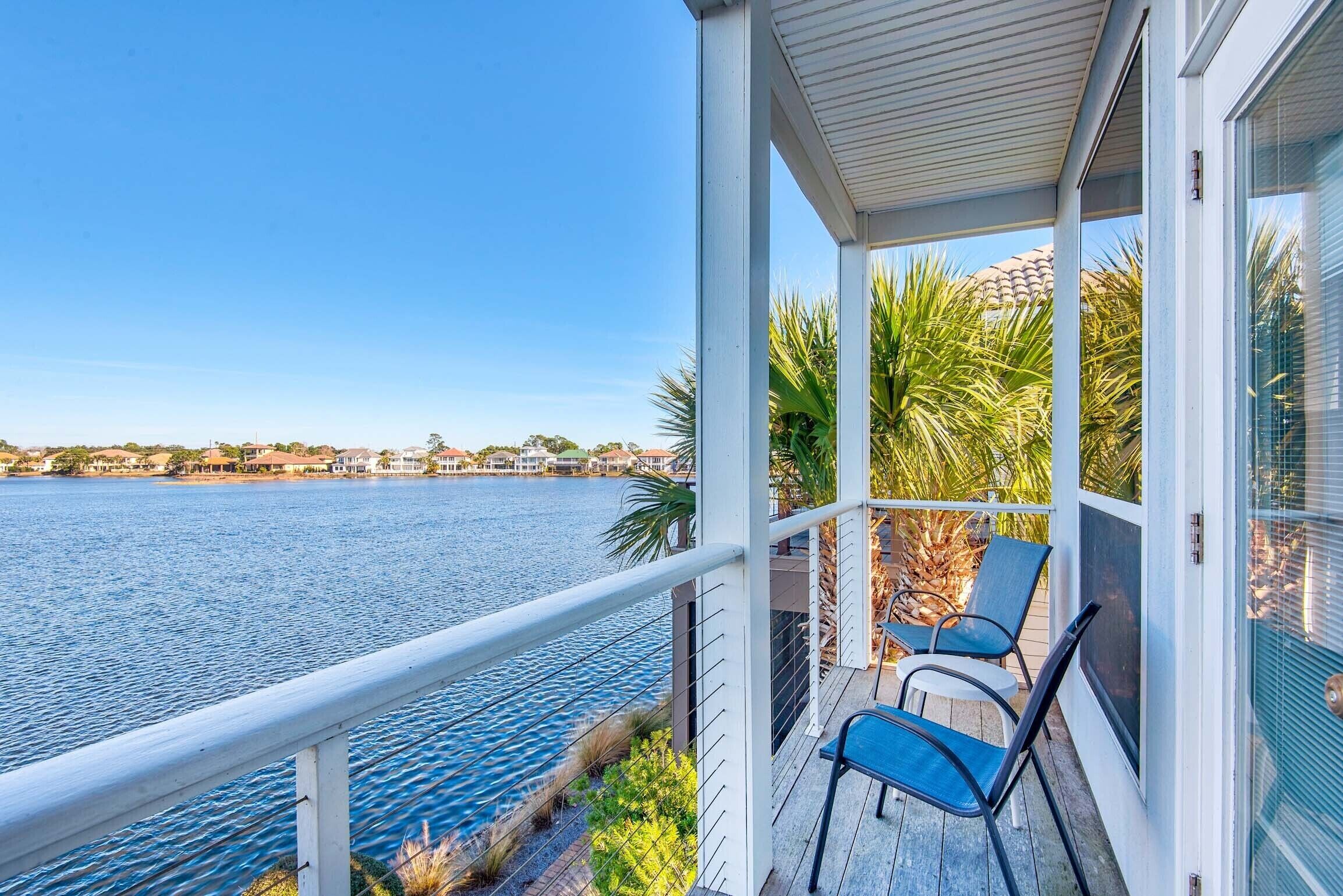 A covered balcony with two chairs and a table, overlooking a calm body of water and a distant shoreline with buildings.