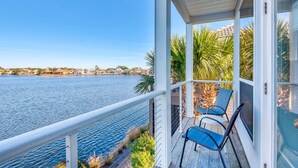A covered balcony with two chairs and a table, overlooking a calm body of water and a distant shoreline with buildings. - ⛱️By Beach-Waterfront Villa- Pool-4BR Destiny Beach Villas 11-Beach Oasis (Destin)