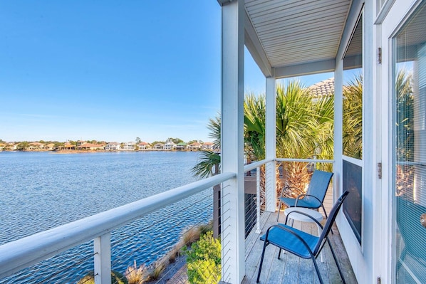 A covered balcony with two chairs and a table, overlooking a calm body of water and a distant shoreline with buildings.