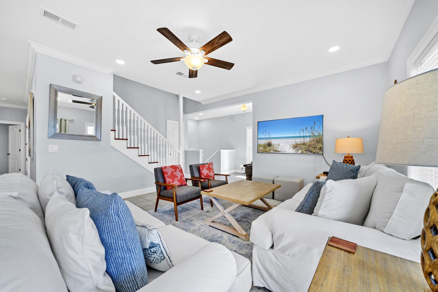 A modern living room with a ceiling fan, a staircase, a beach-themed wall art, and a dining area.