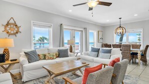 A spacious living room with a white sofa, a wooden coffee table, and wicker chairs. There is a ceiling fan, large windows with a view, and a dining area with wicker furniture. - 🌊3 Generations Beach House | Beachfront | Private Pool | Sleeps 17 (Santa Rosa Beach)