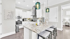 Una cocina moderna con armarios blancos, una isla de granito, electrodomésticos de acero inoxidable e iluminación colgante.