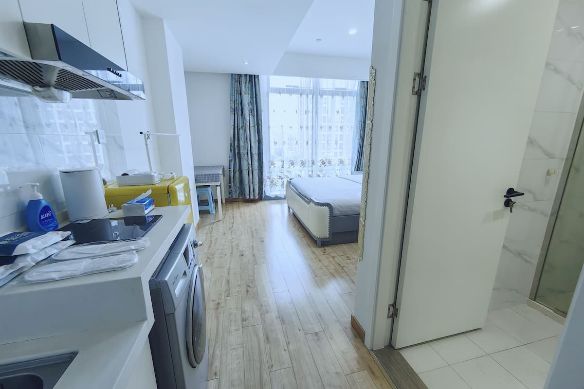 A hotel room with a washing machine, a small table with a chair, a bed, and a bathroom with a glass door.