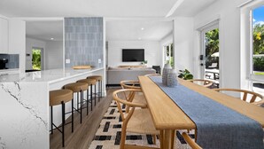 A modern kitchen with a marble countertop and bar stools, a dining area with a wooden table and chairs, and a living area with a sofa and TV.