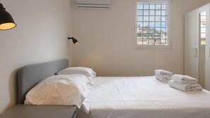 Cama bem arrumada com lençóis brancos e toalhas dobradas, luminária de cabeceira e unidade de ar condicionado na parede.