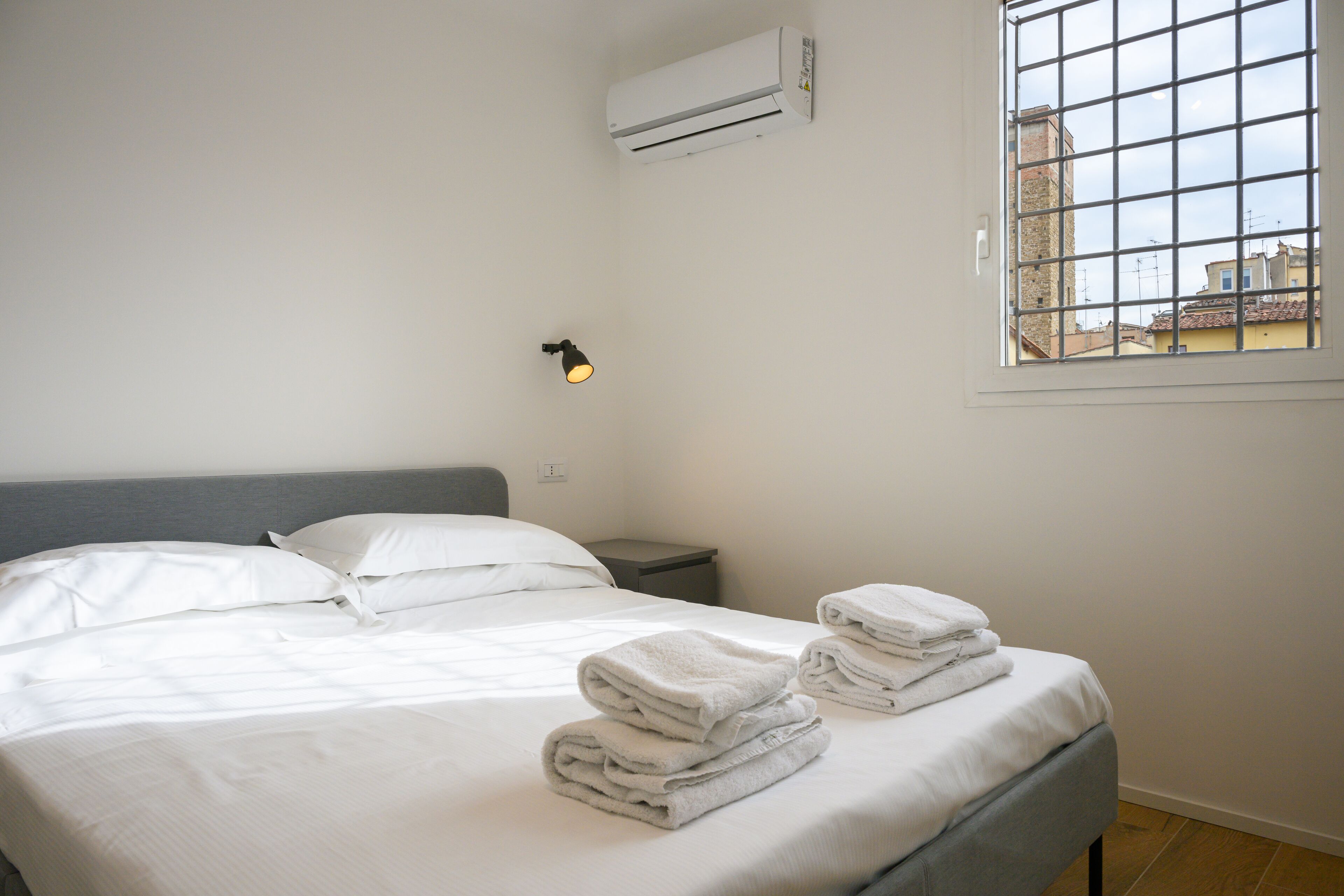 Une chambre d’hôtel comprenant un lit, une table de chevet, un climatiseur, une fenêtre donnant sur des immeubles et un lampadaire fixé au mur.