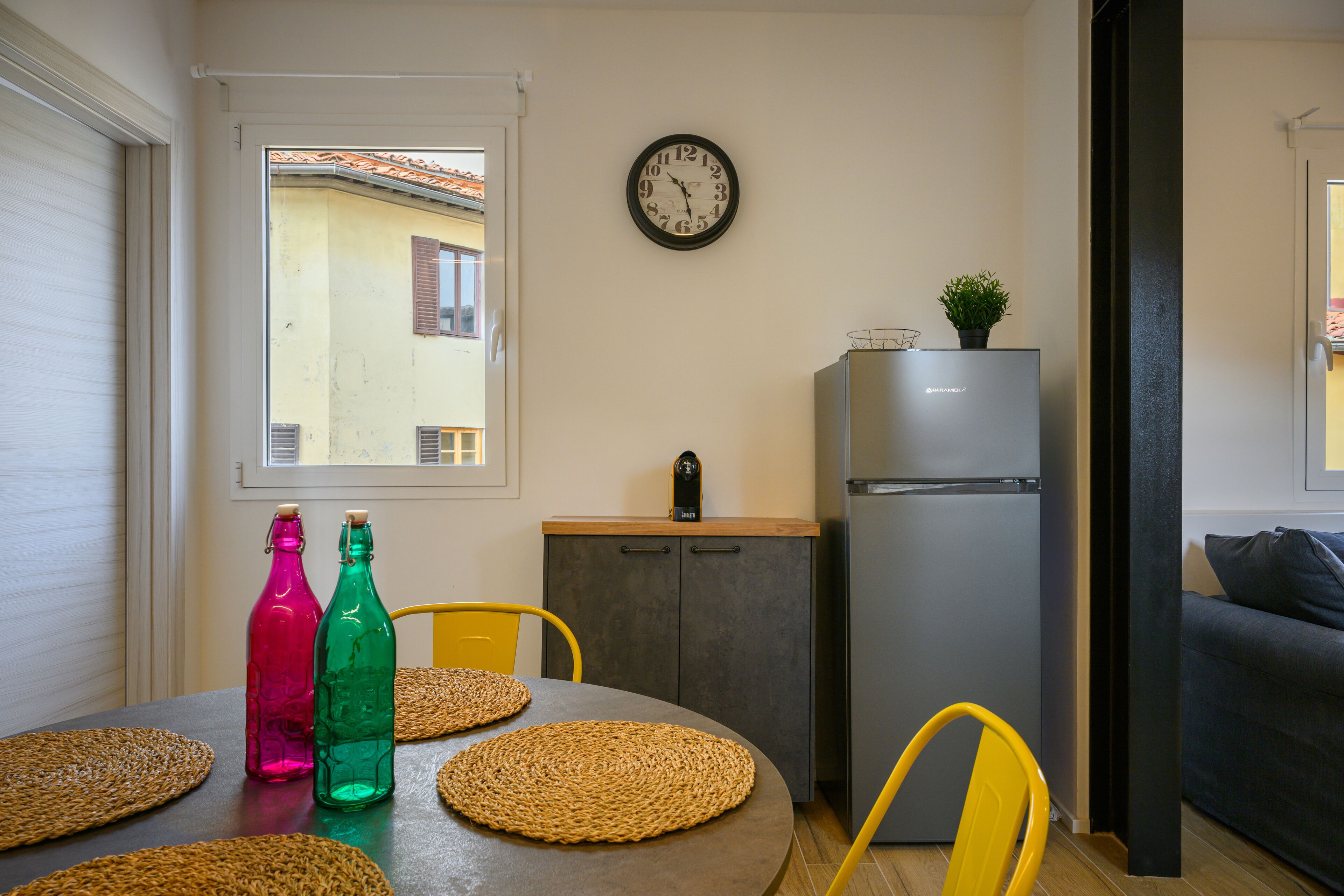 A compact kitchen with a refrigerator, a clock, and a window with a view of buildings.