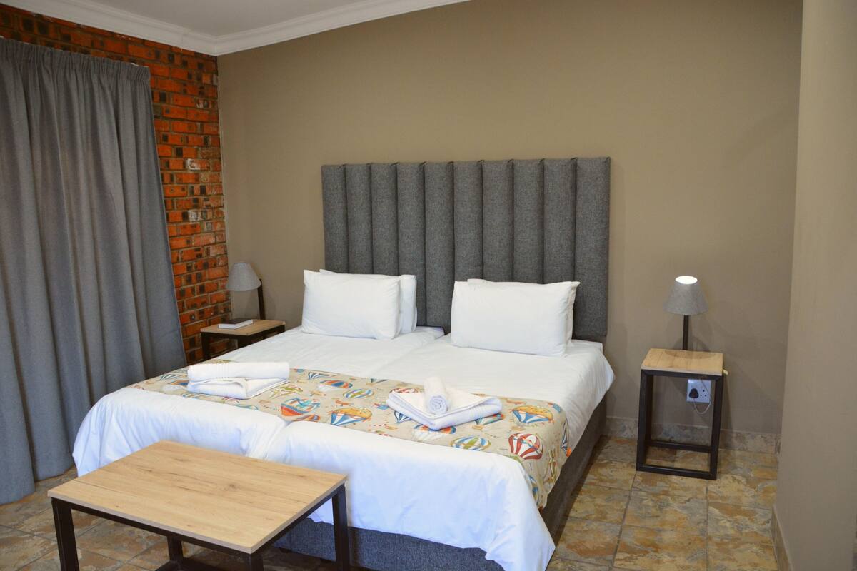 Een hotelkamer met een bed, twee kussens, een nachtkastje met een lamp, een houten tafel en een bakstenen muur.