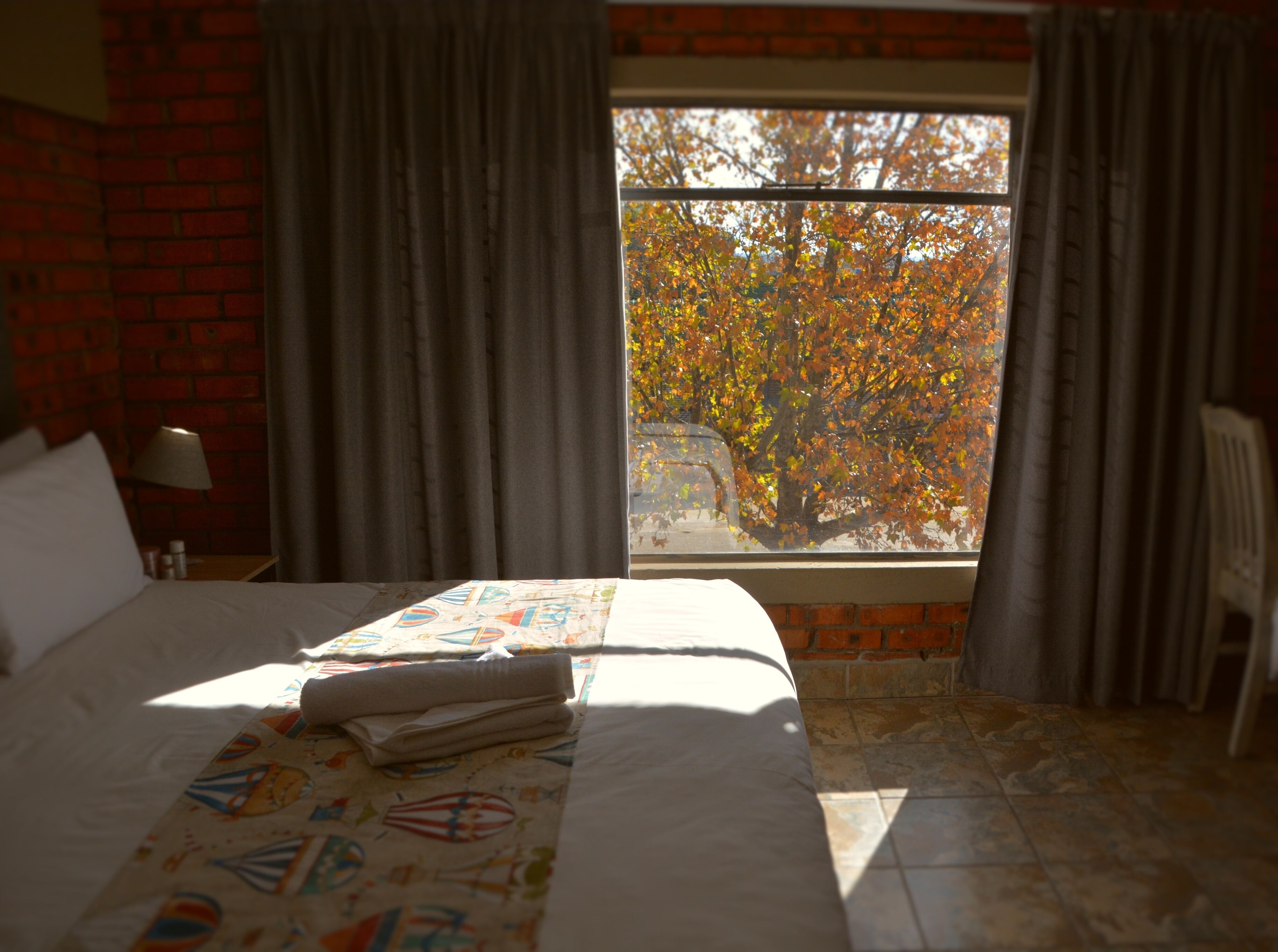 A hotel room with a brick wall, a large window with curtains, a bed with a colorful bedspread, and a chair.