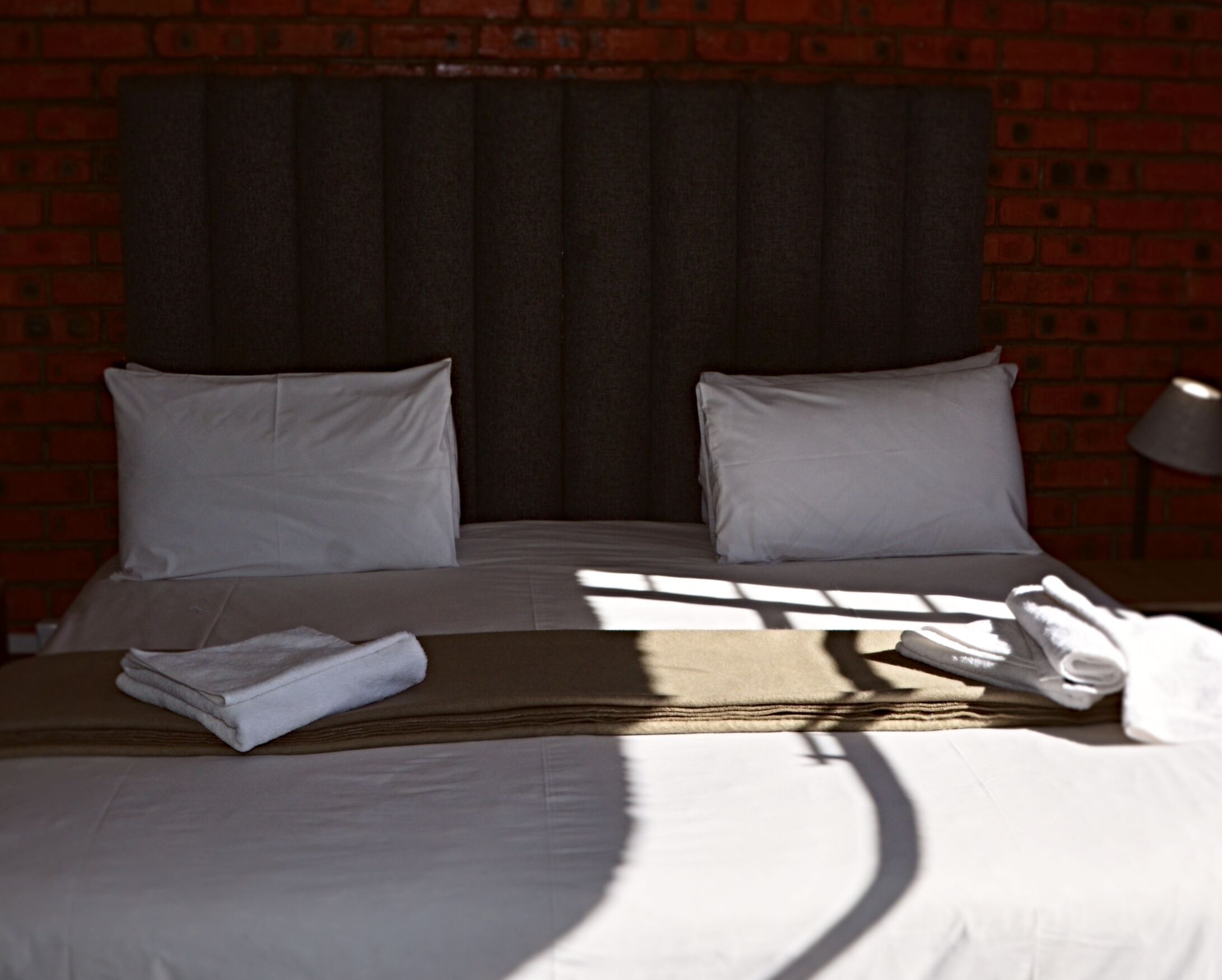 A neatly made bed with white linens and a folded towel, set against a brick wall with a dark headboard.