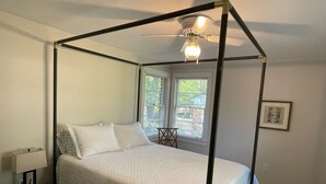 A four-poster bed with a white bedspread and pillows, a bedside table with a lamp, and a ceiling fan.