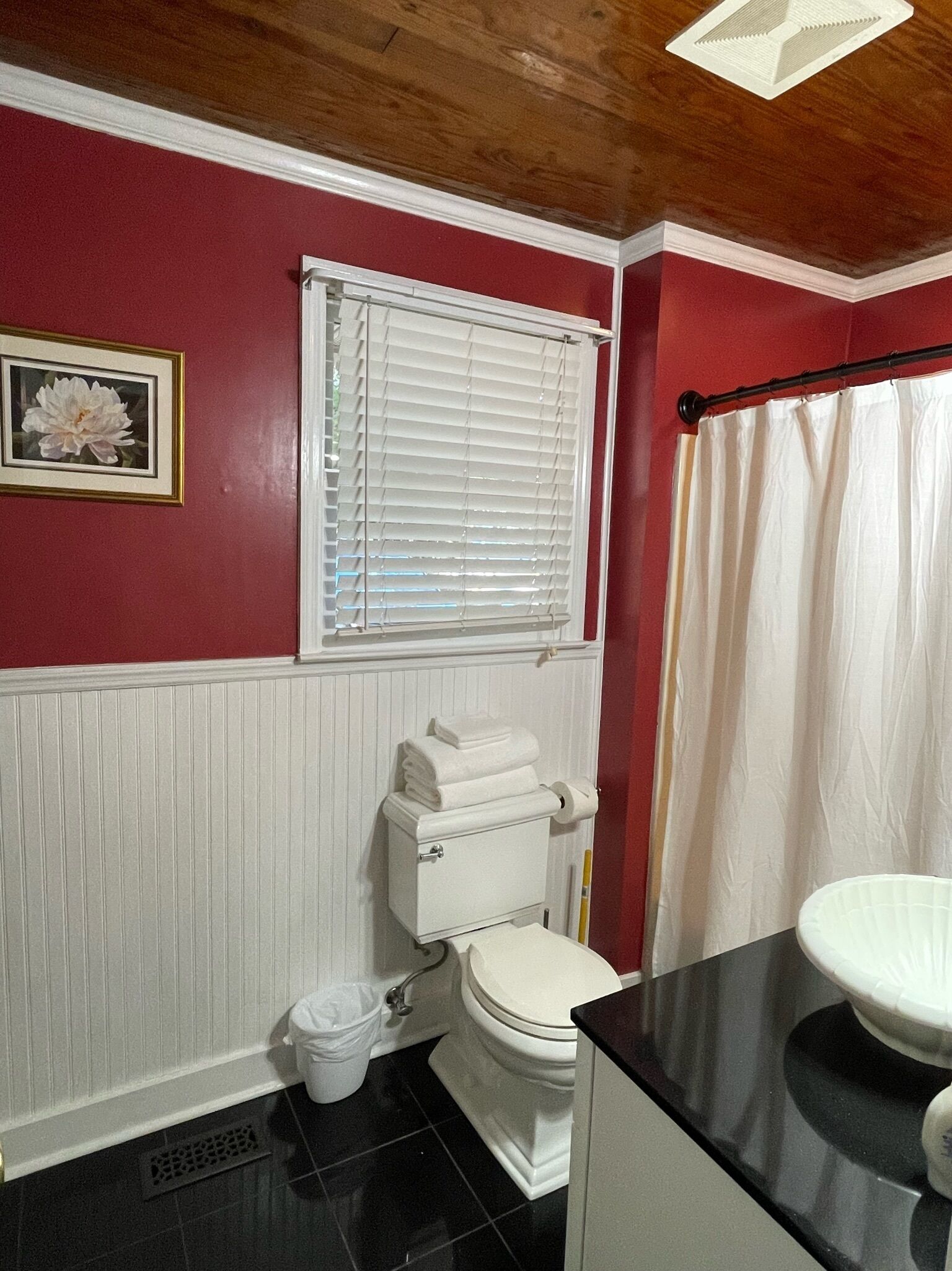 Ein Badezimmer mit Toilette, Waschbecken, einem Fenster mit Jalousien, einer Duschvorhang und einem gerahmten Bild an der Wand.